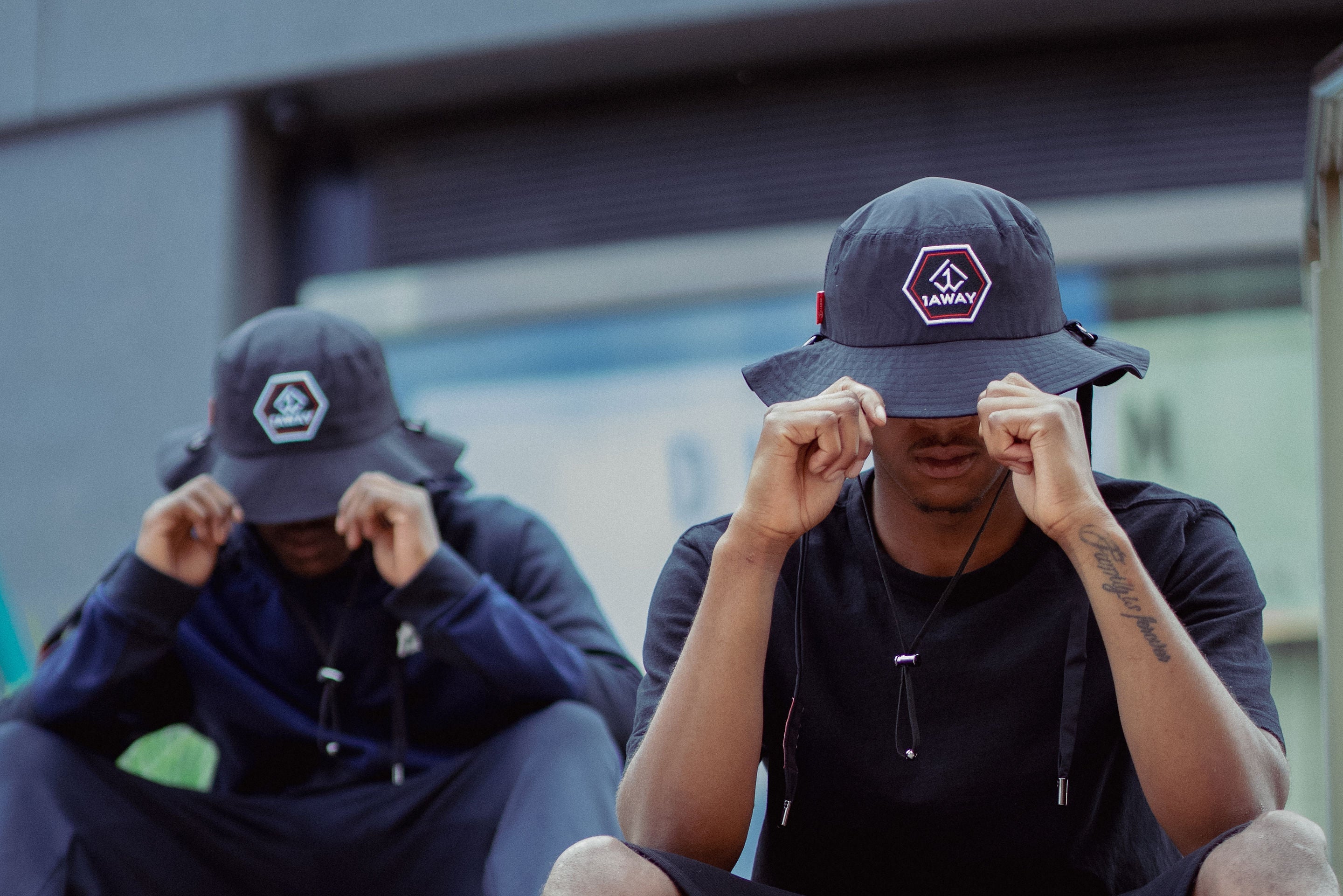 Two people sitting on steps wearing bucket hats with visible 1AWAY brand logos.