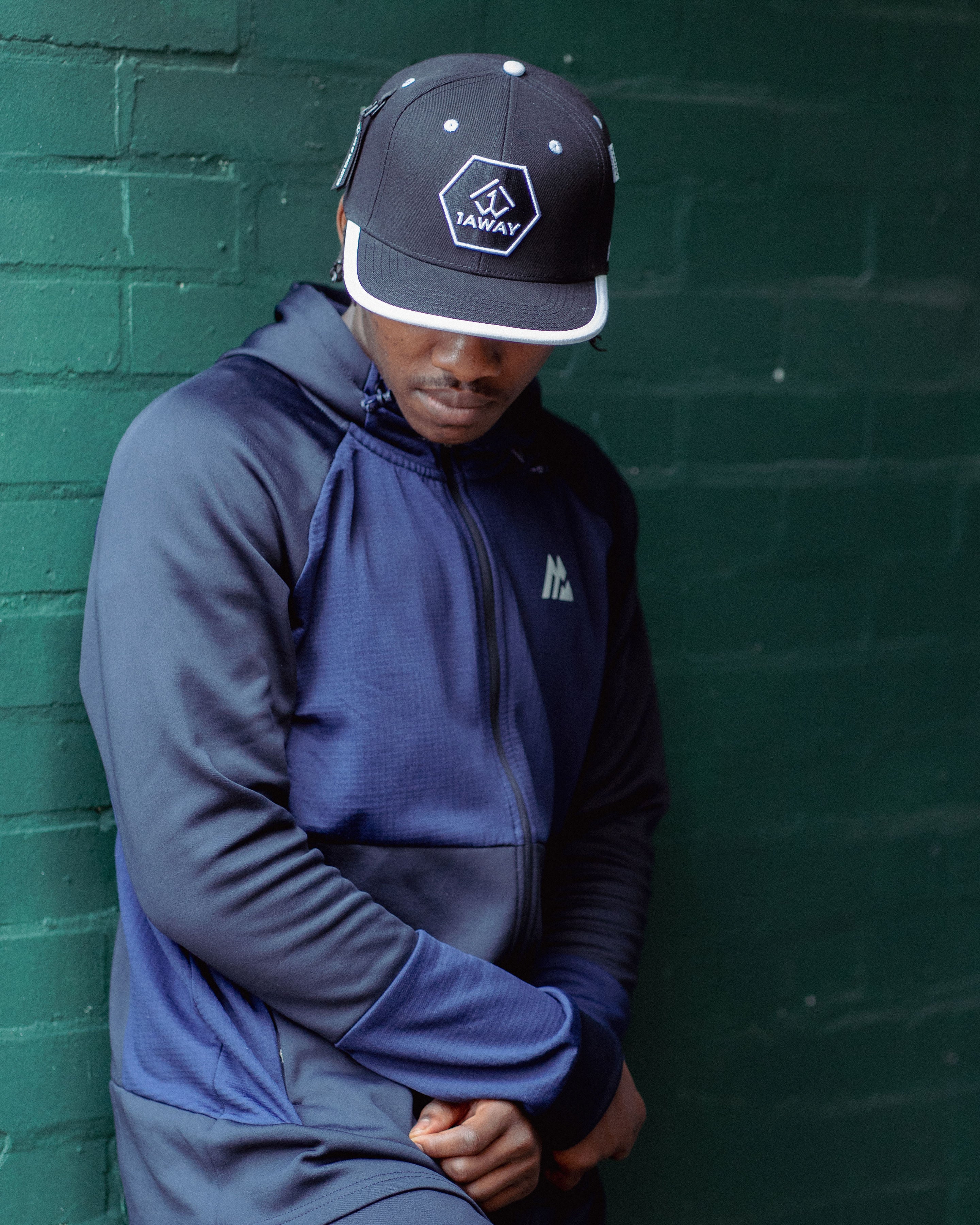 Person wearing a blue tracksuit and black 1AWAY 0001 snapback with 1AWAY logo against a green brick wall
