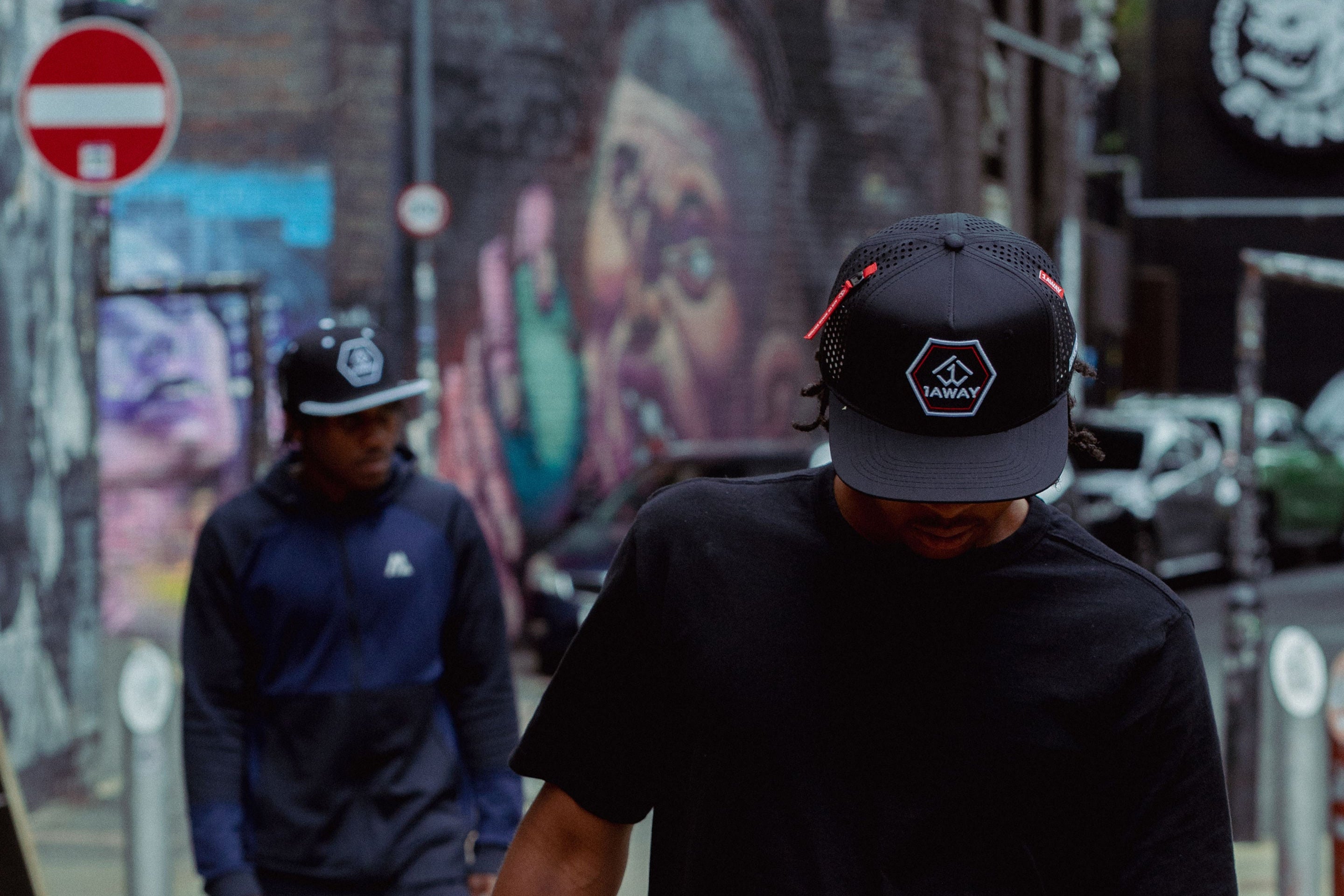 Two individuals walking on a street with graffiti art in the background, wearing 1AWAY snapbacks 