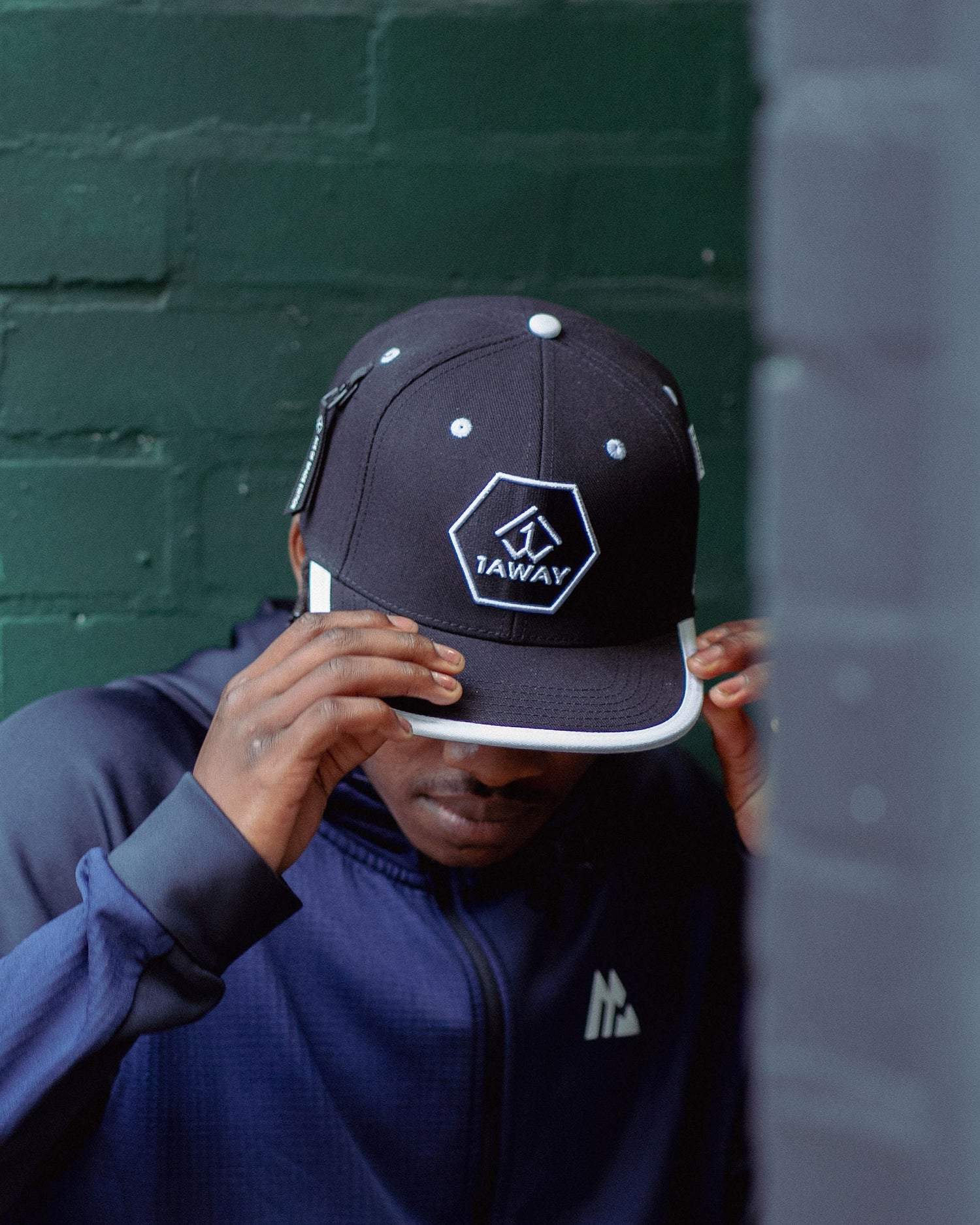 Young black man wearing a dark blue jacket and black 0001 1Away snapback cap with a logo against a green wall.