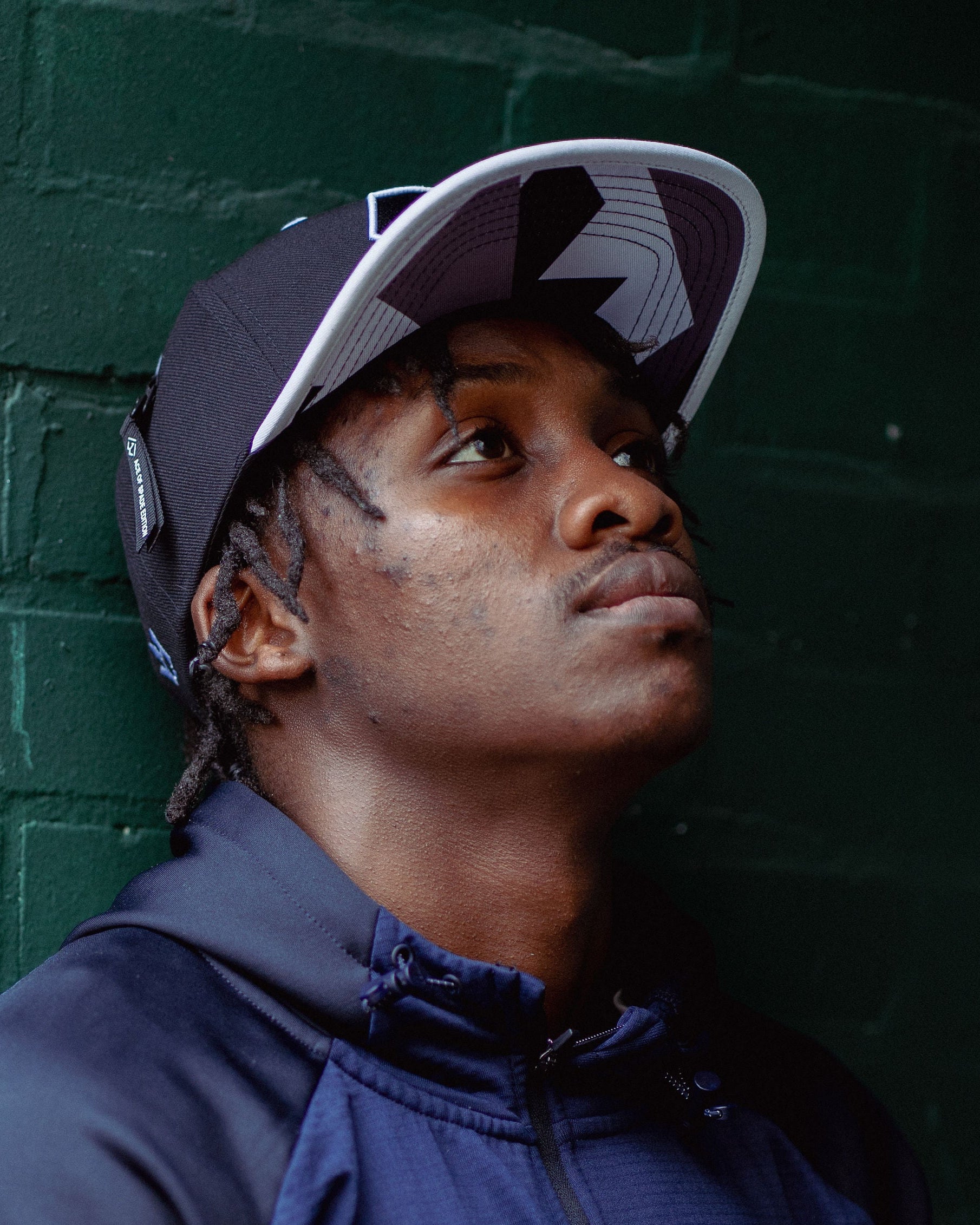Person wearing a blue jacket and 1AWAY snapback cap against a green brick wall