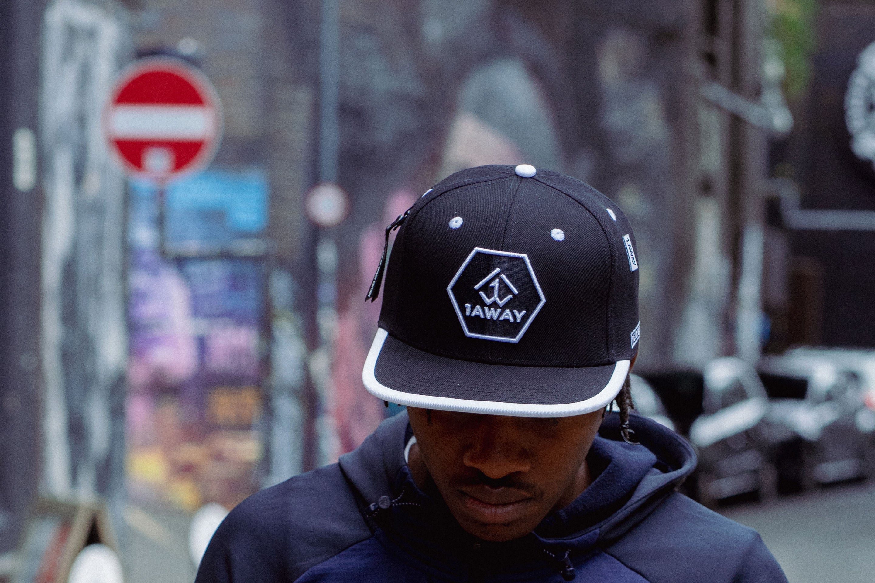 Person wearing a navy jacket and black cap with a logo, standing on a street.