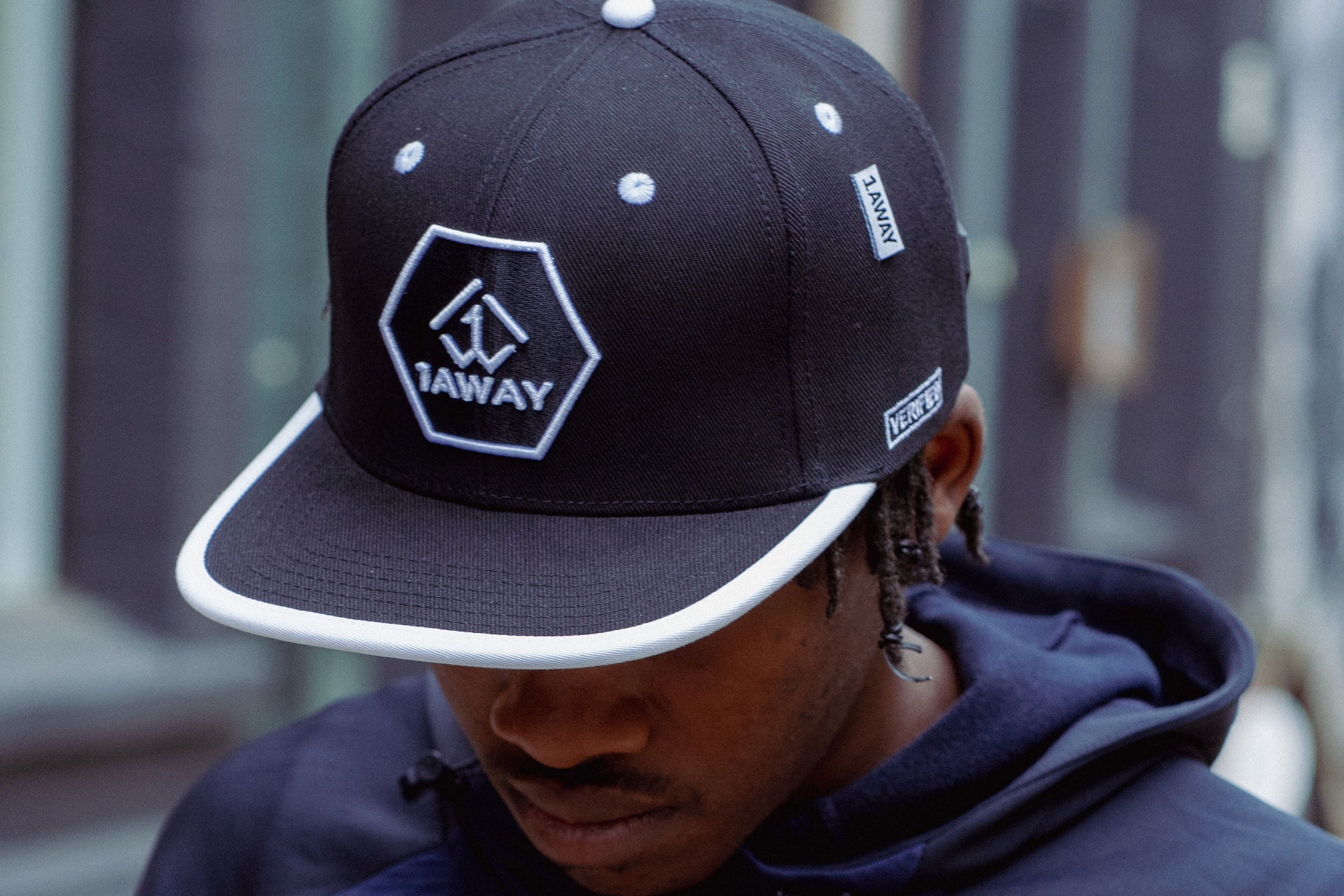 Young black man in the UK wearing a black a 1AWAY snapback cap with a logo and a navy hoodie