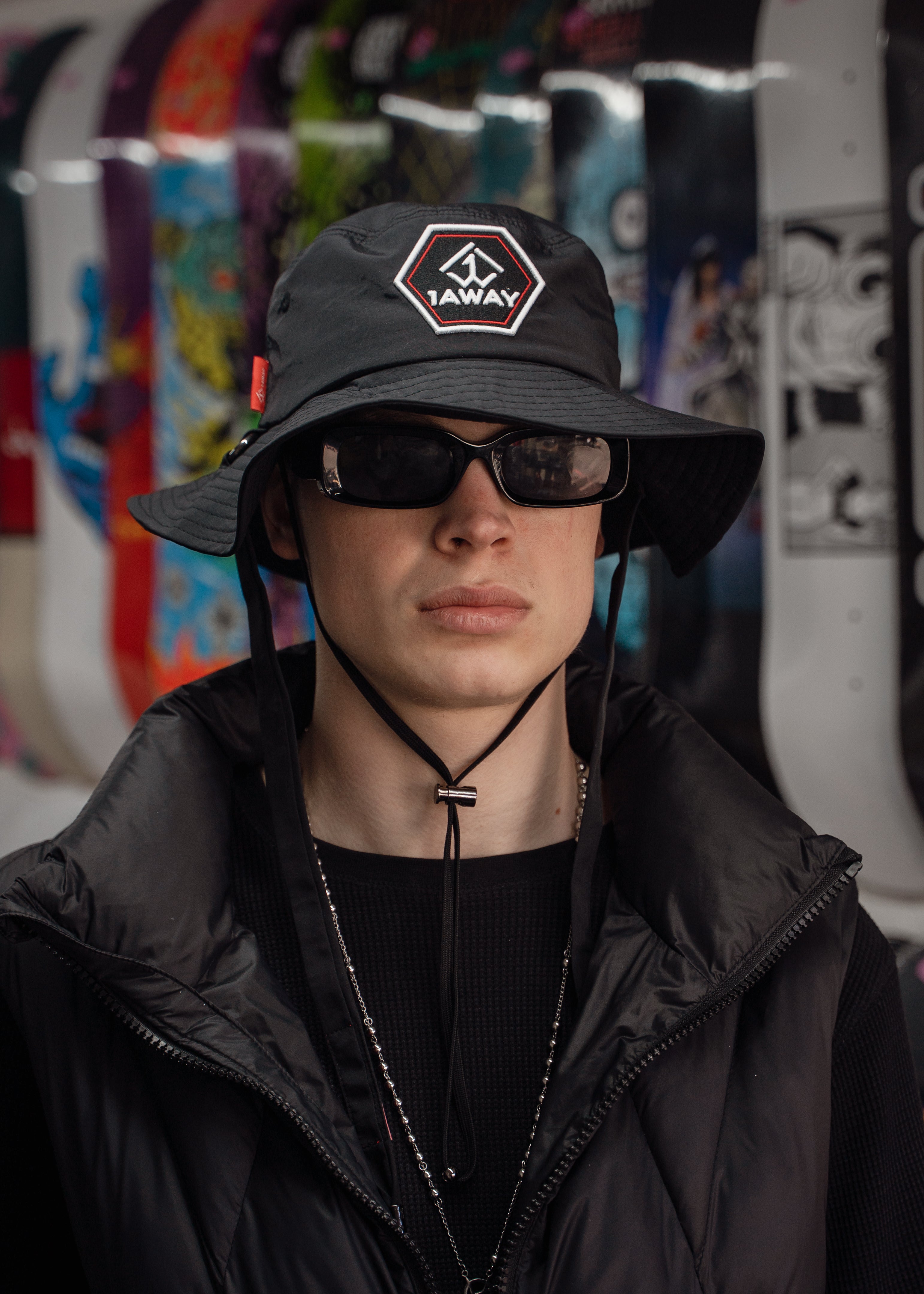 Person wearing a black bucket hat with a logo and sunglasses, standing in front of colorful skateboards.