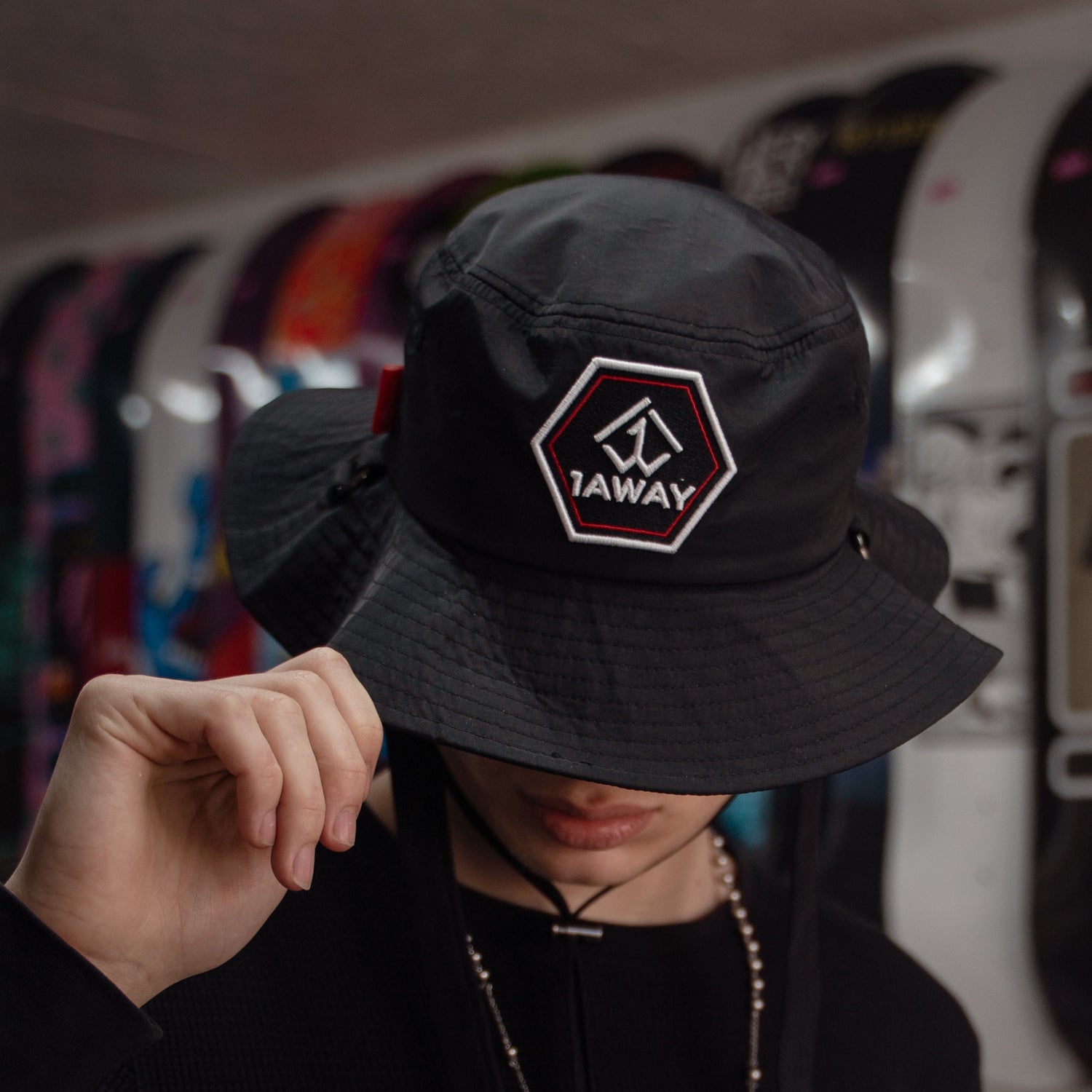 Person wearing a black 1AWAY bucket hat with a logo in a skate shop with colourful skateboards in the background