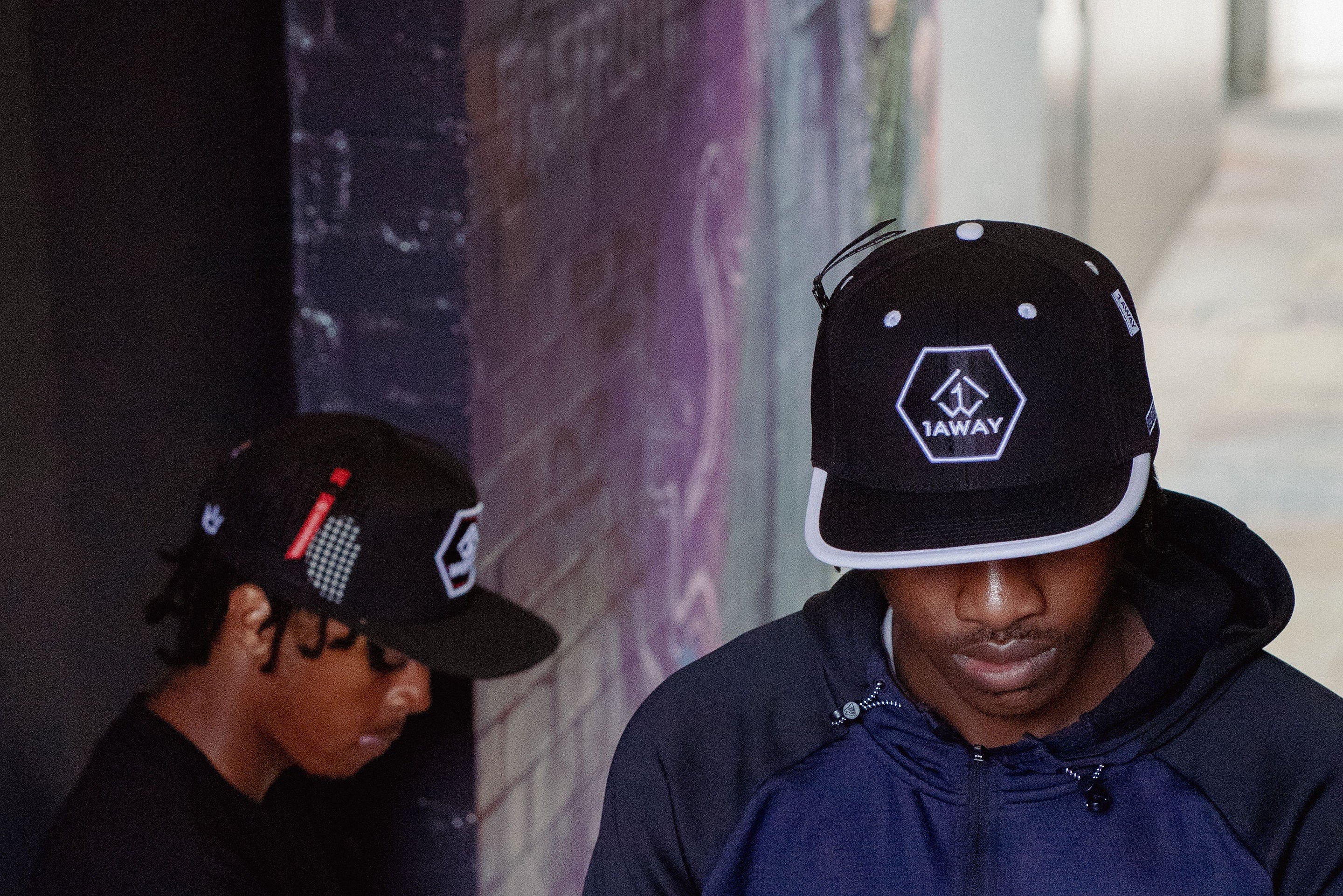 Two individuals wearing black caps with logos, sitting against a wall with graffiti, wearing 1AWAY snapbacks
