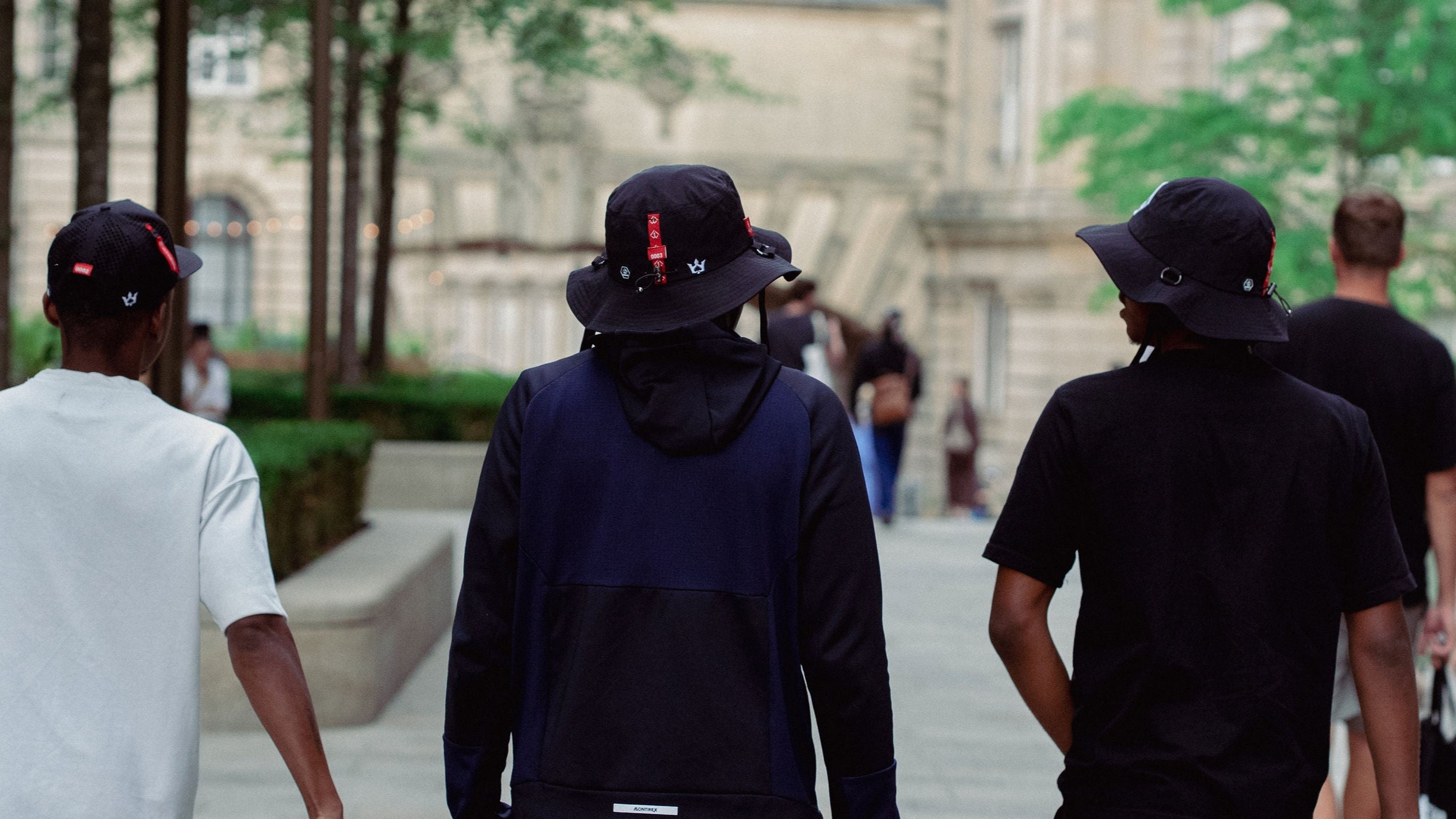 Three people walking on a city street, wearing dark hats and hoodies, wearing 1AWAY hats